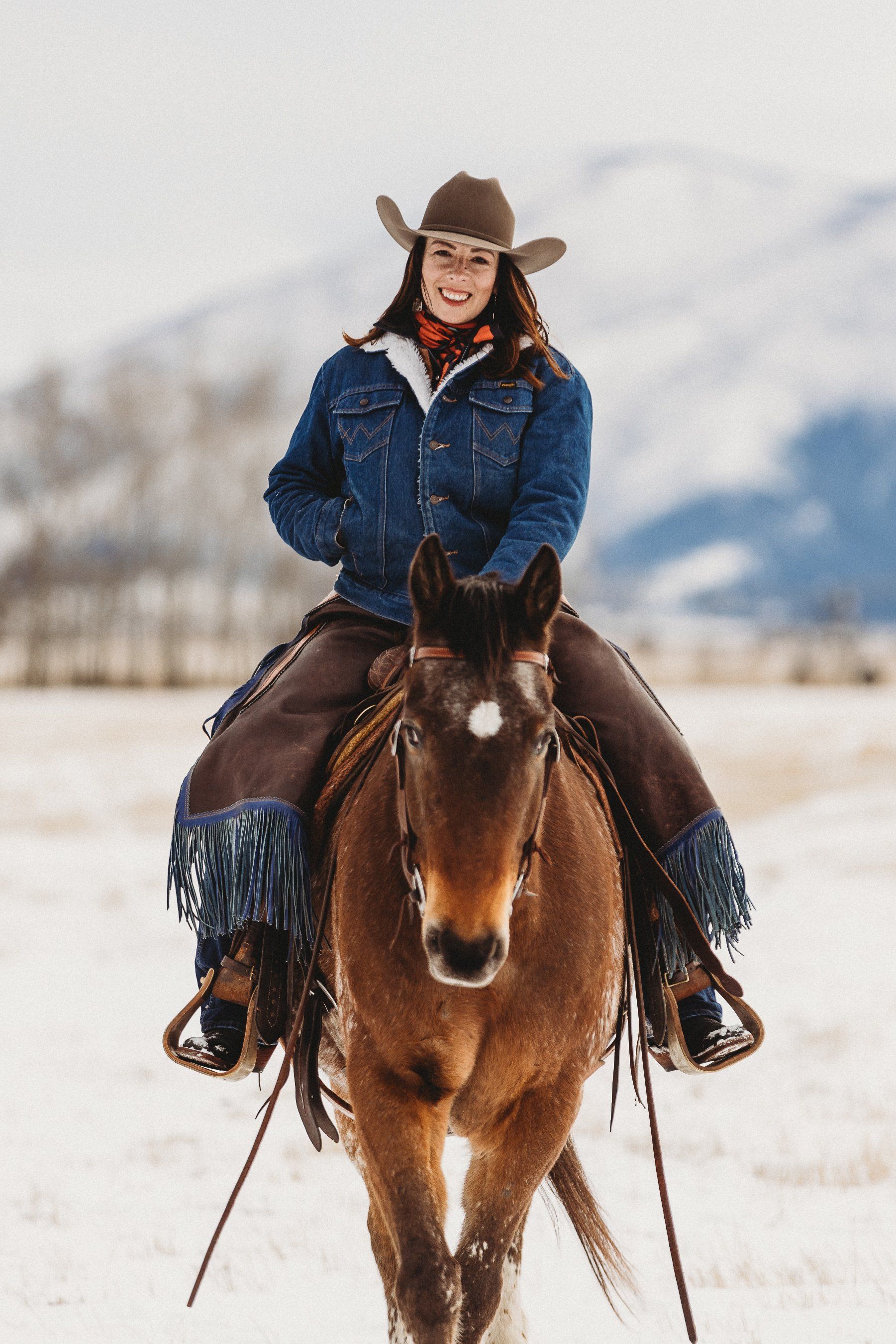 Woman on a brown horse, wearing a cowboy hat and denim jacket, riding in snowy field.