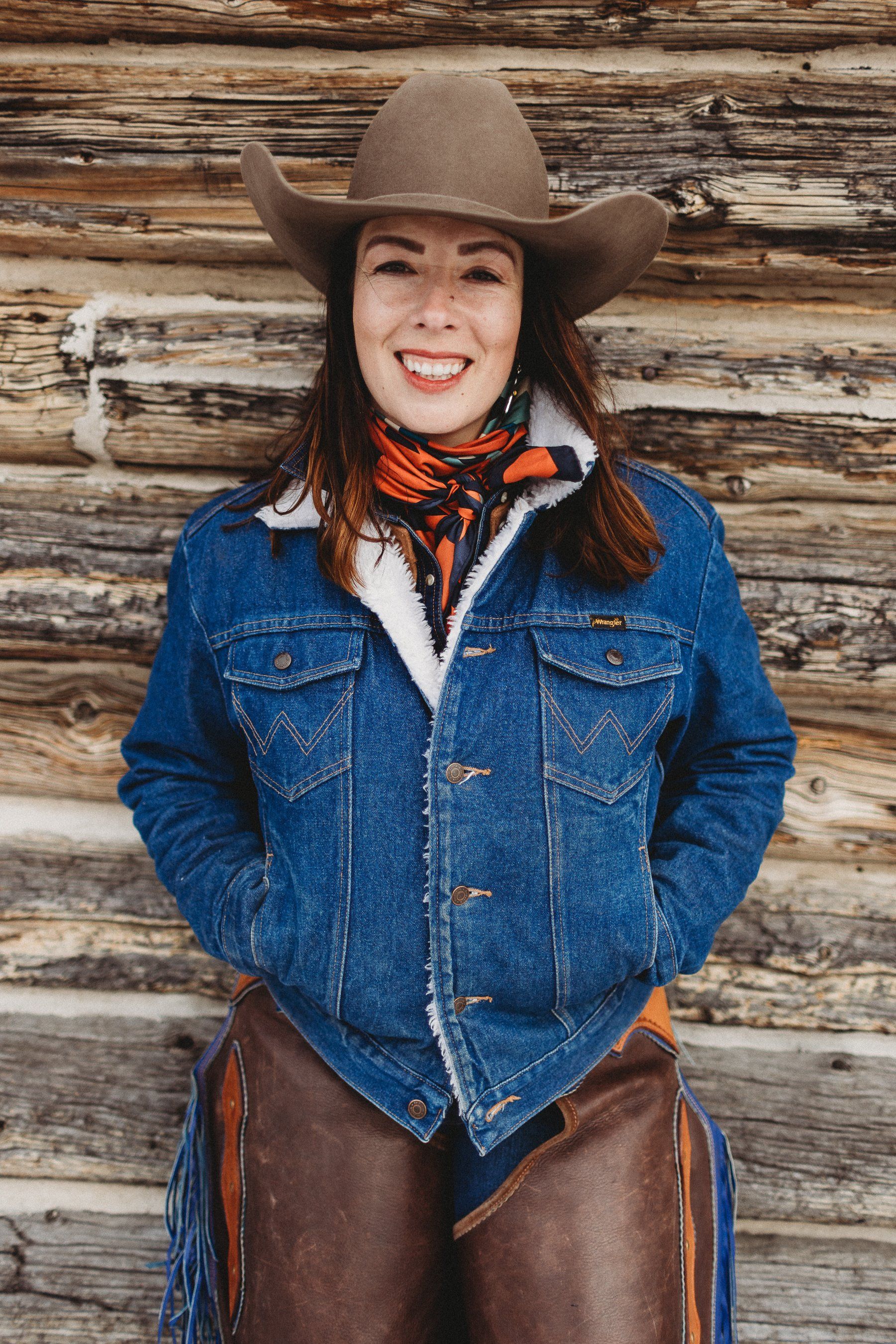 Cowgirl in a denim jacket and cowboy hat smiling, leaning against a wooden wall.