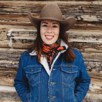 Woman in cowboy hat and denim jacket smiles against a wood-paneled wall.