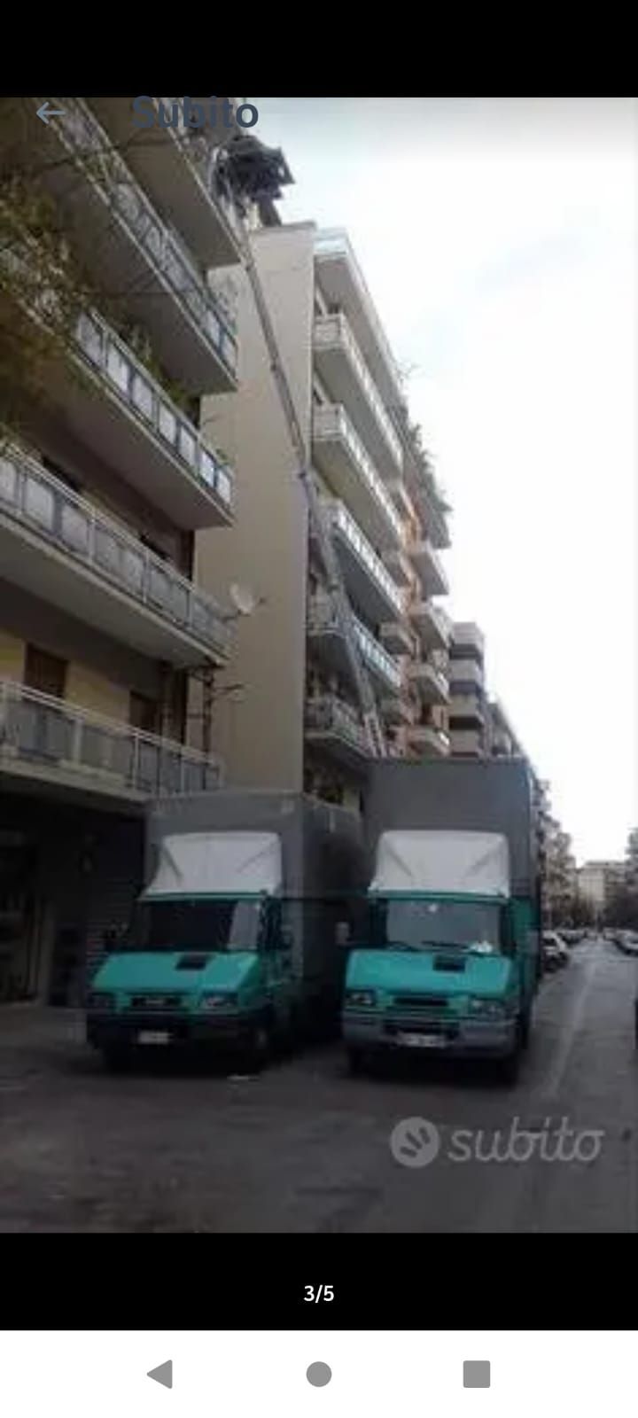 Due camion per traslochi color verde acqua parcheggiati davanti a un alto condominio.