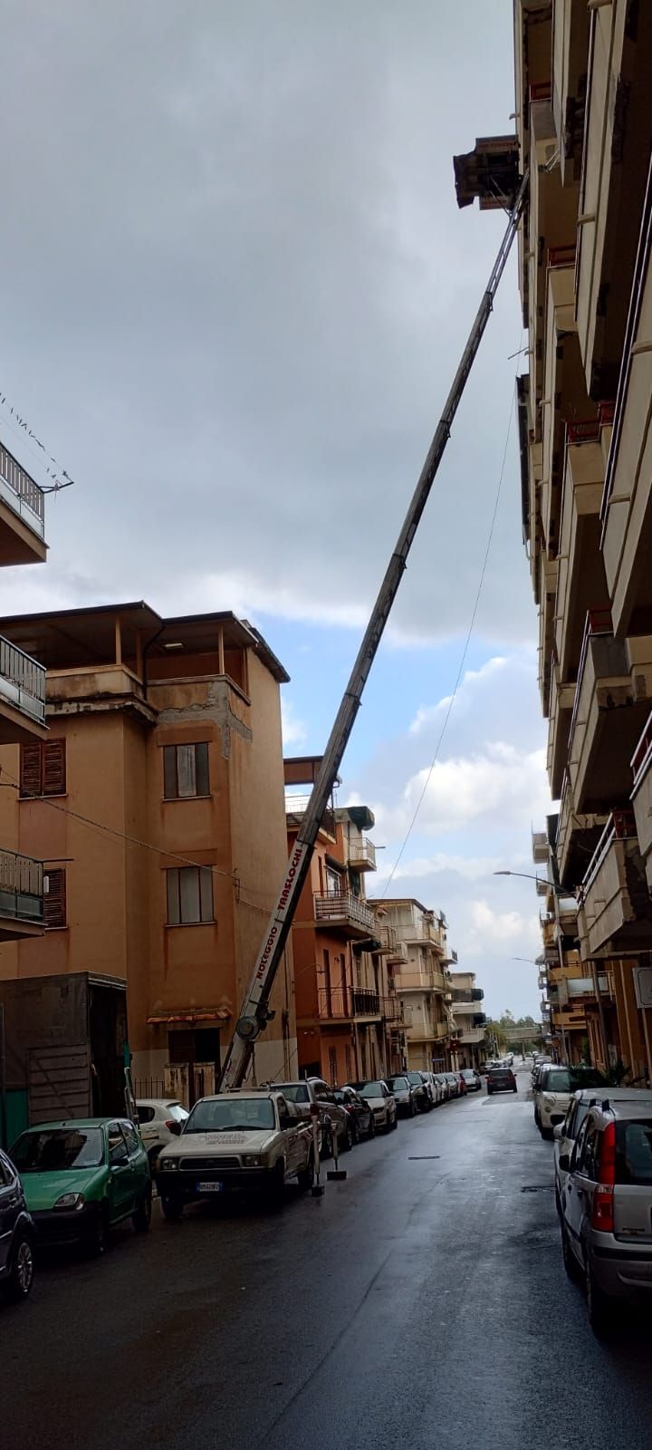 Una gru a torre raggiunge un alto condominio in una strada stretta. Le auto sono parcheggiate su entrambi i lati della strada.