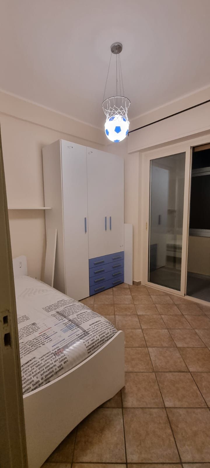 Camera da letto con mobili bianchi, un letto, un grande armadio, una porta finestra e una lampada a sospensione floreale.