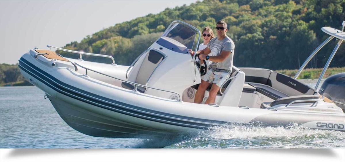 Two people riding a white speedboat on blue water with a green, hilly shoreline in the background