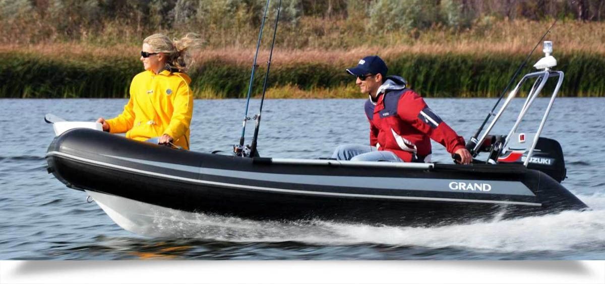 Two people riding in a black motorboat on a lake, with fishing rods and reeds in the background