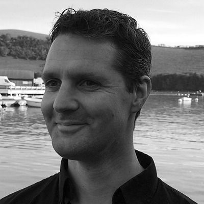 Man smiling in a black shirt by the water, with boats and hills in the background