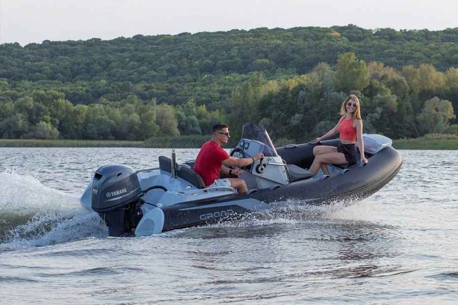 Three people ride a gray motorboat on a lake, with a forested shoreline in the background.