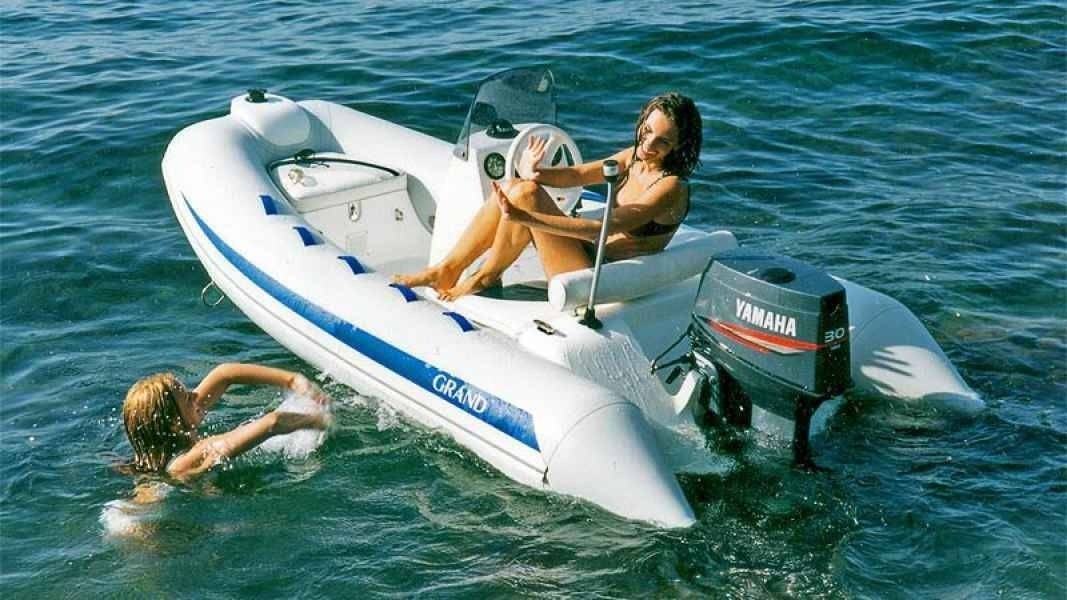 Two people by a small white motorboat on blue water, one sitting and one swimming beside it