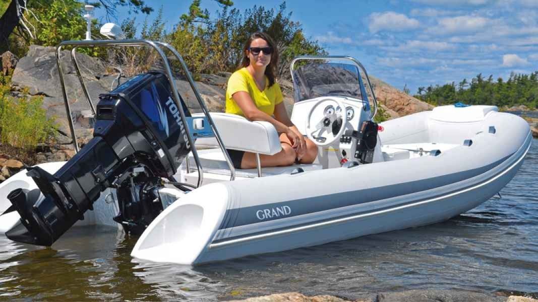 Woman in a yellow shirt sits in a white inflatable Grand Silver Line 470 motorboat on a sunny beach shore.