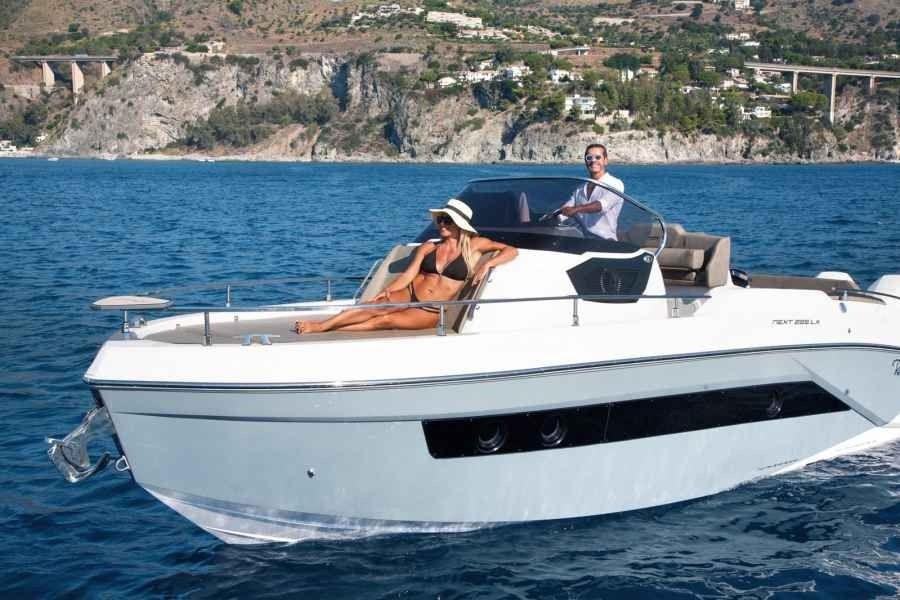 Two people on a white motorboat cruising on blue water near a rocky coastline.
