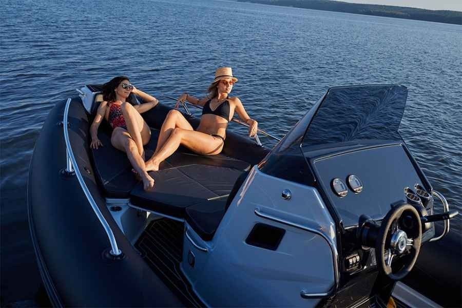 Two people lounging on a sleek blue motorboat on open water, one steering with a drink in hand.