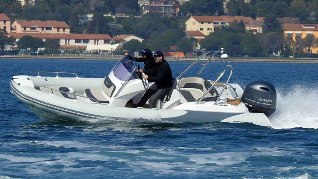 Two people ride a white grand golden line 580 speedboat across choppy blue water with a shoreline town in the background