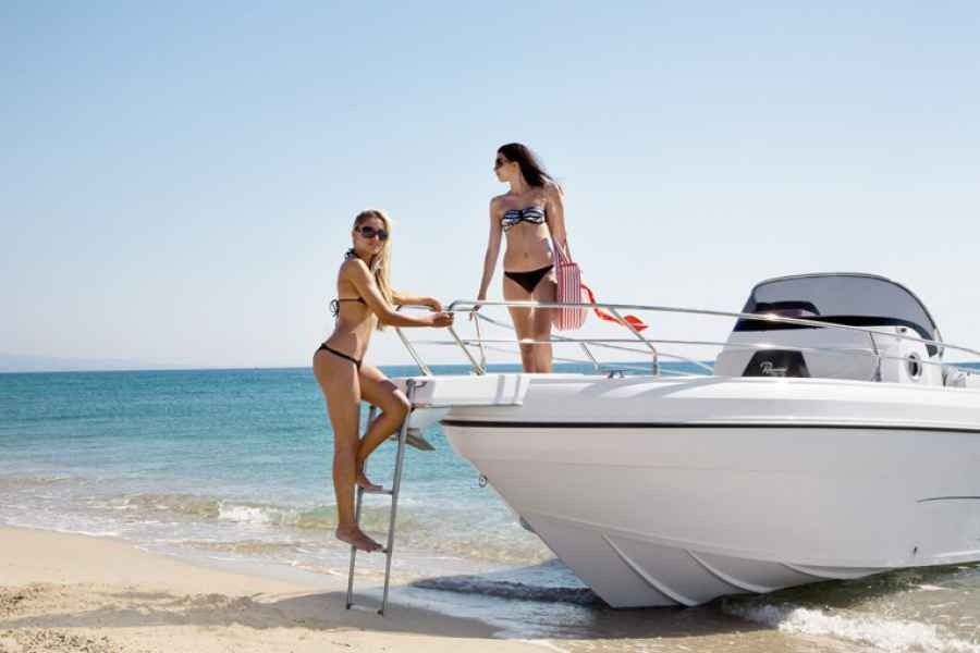 Two women on a white boat at a sunny beach, one climbing aboard from the water.