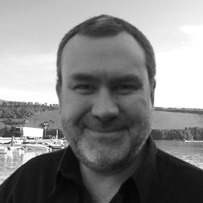 Smiling man in a dark shirt standing outdoors by the water with hills in the background