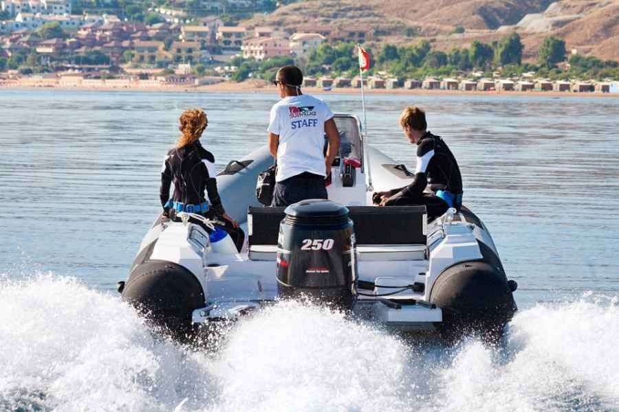 Three people ride a boat on calm water, with a shoreline town in the background.