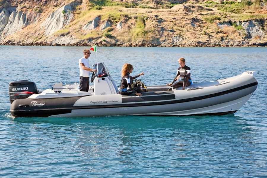 People riding in a gray inflatable ranieri cayman 23 sport diving rib motorboat on calm blue water near a rocky shoreline