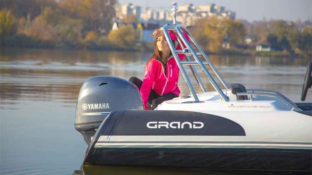 Woman in a bright pink jacket seated on a grand golden line 500 boat by calm water, with a ladder and distant shoreline.