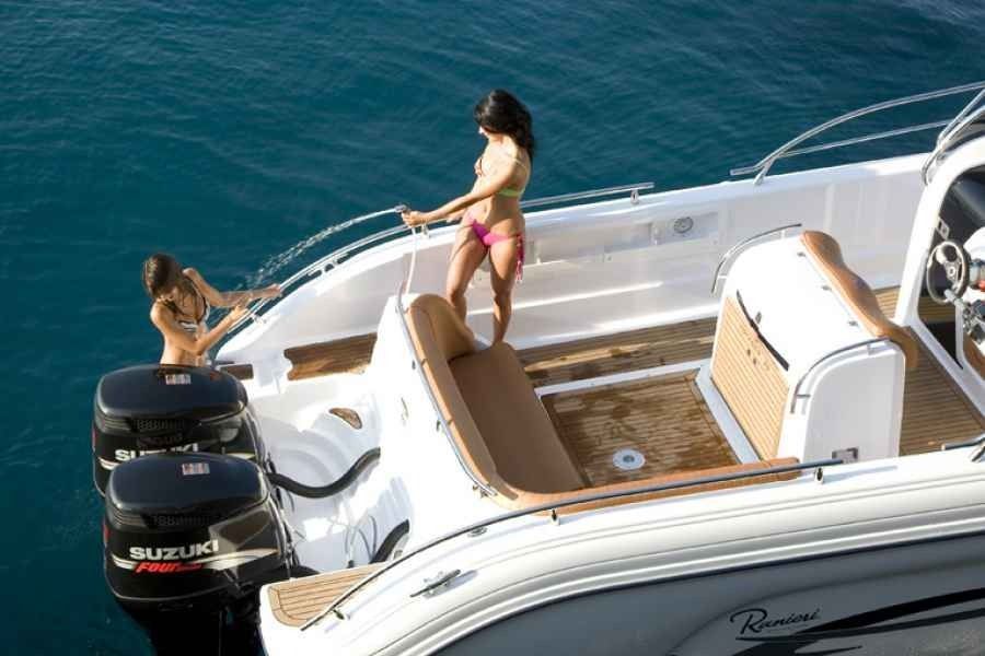 Two people on a white boat in blue water, one standing on deck and one in the sea with an outboard motor visible