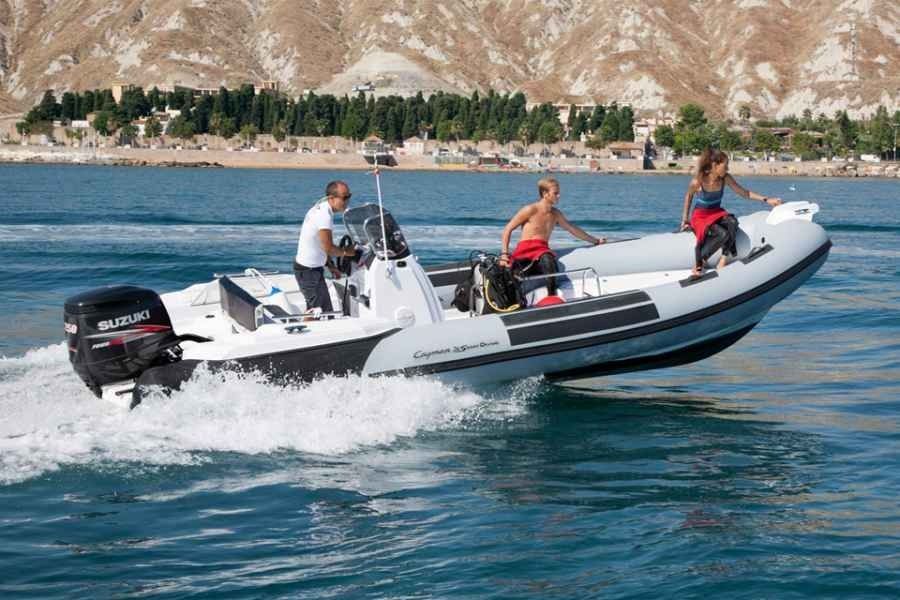 Three people ride a white ranieri cayman 26 sport diving rib speedboat cutting across blue water near a rocky shoreline.