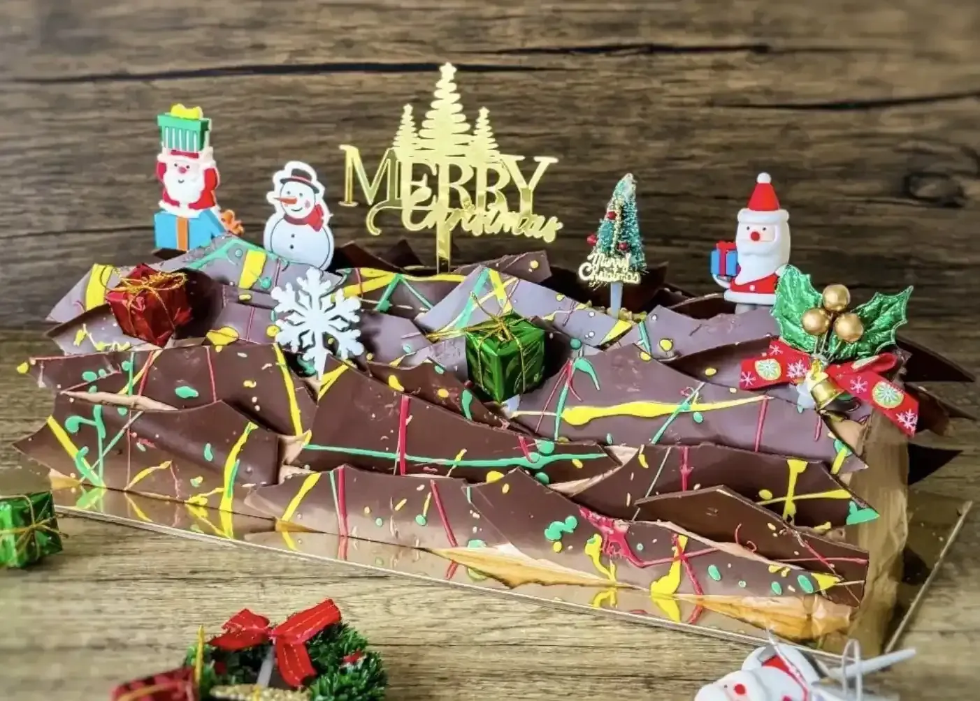 a christmas chocolate log cake decorated with crispy chocolate flakes, ornaments of santa claus, snow men, and christmas trees