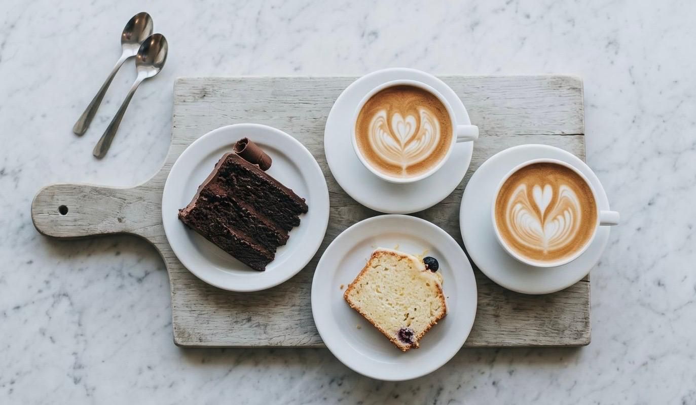 An overhead flat lay photograph of a wooden serving board on a marble countertop, featuring two plates with slices of chocolate cake and lemon loaf, alongside two cups of latte with latte art and two small spoons.