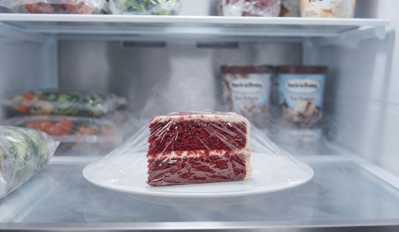 A neat slice of red velvet cake, wrapped in clear plastic film, sitting on a white ceramic plate on a clean freezer shelf, surrounded by cold air vapor and other frozen items like ice cream pints.