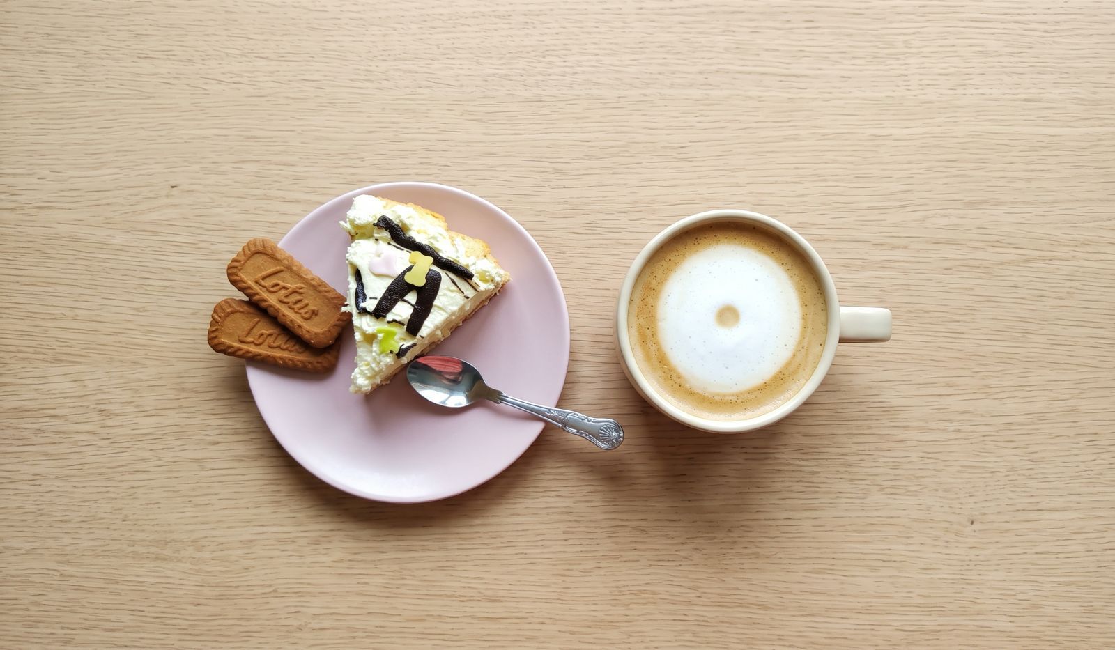 An overhead view of a small pink plate holding a decorative slice of layered cake, two Lotus Biscoff cookies, and a patterned silver spoon, set next to a ceramic cup of coffee with a circular foam pattern, all on a light wooden table surface.