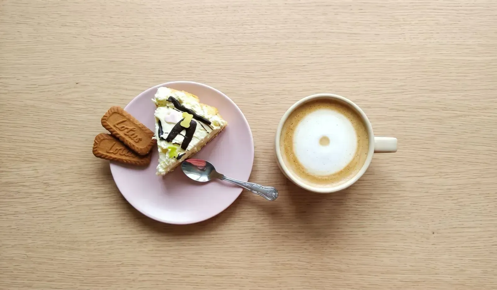 An overhead view of a small pink plate holding a decorative slice of layered cake, two Lotus Biscoff cookies, and a patterned silver spoon, set next to a ceramic cup of coffee with a circular foam pattern, all on a light wooden table surface.