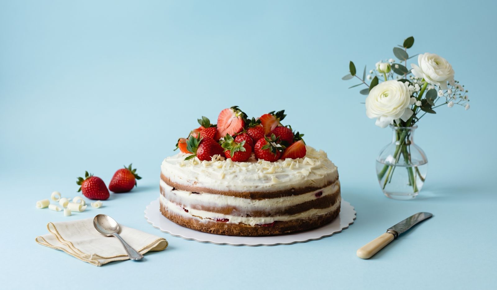 a gluten free strawberry cake placed in on a clean cotton blue backdraft. decorated with strawberries, utensils, and vased plants