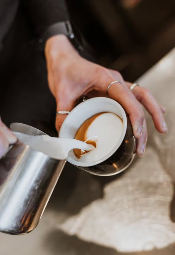 Persona che versa latte montato a vapore in una tazzina di caffè espresso, creando una sorta di latte art.