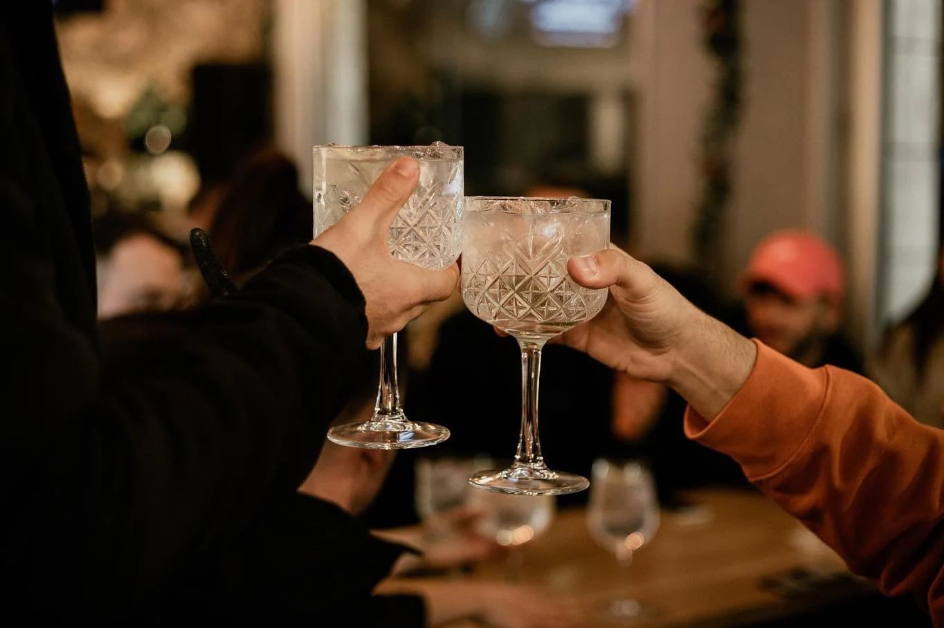 Due persone brindano con dei bicchieri di gin decorati in un bar scarsamente illuminato, festeggiando con altre persone che si intravedono sullo sfondo.