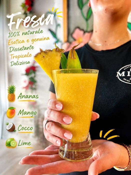 Mano che tiene un drink tropicale giallo con una fetta di ananas come decorazione, accanto a un menu di succhi freschi