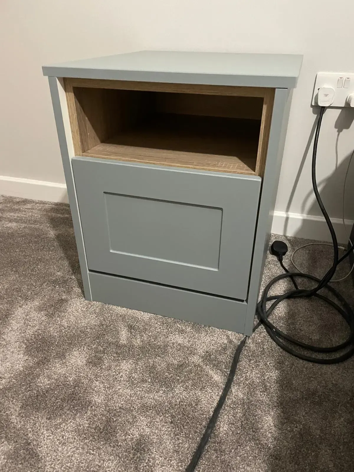 Gray cabinet with open wooden shelf, sitting on carpet beside a wall outlet and cords.