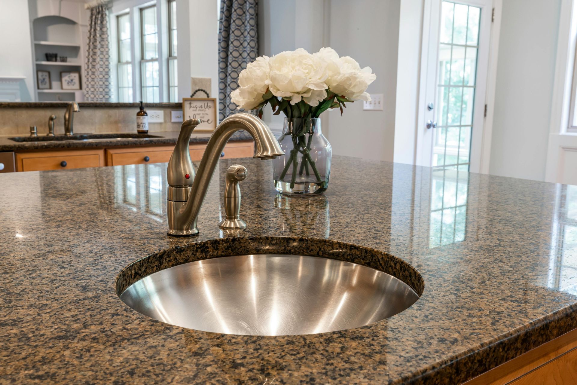 Kitchen island with granite countertop, stainless steel sink, and vase of white flowers near bright windows