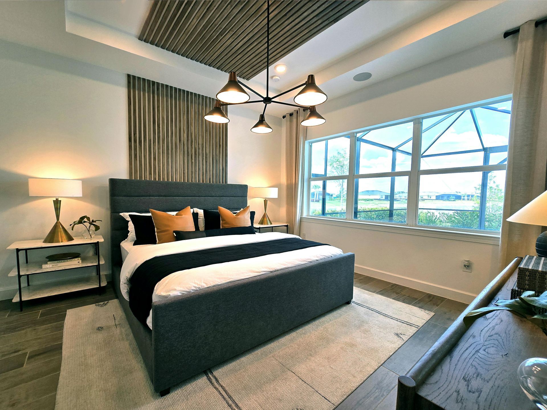 A modern bedroom with a dark gray upholstered bed, wooden wall art, a black metal chandelier, and a large window.
