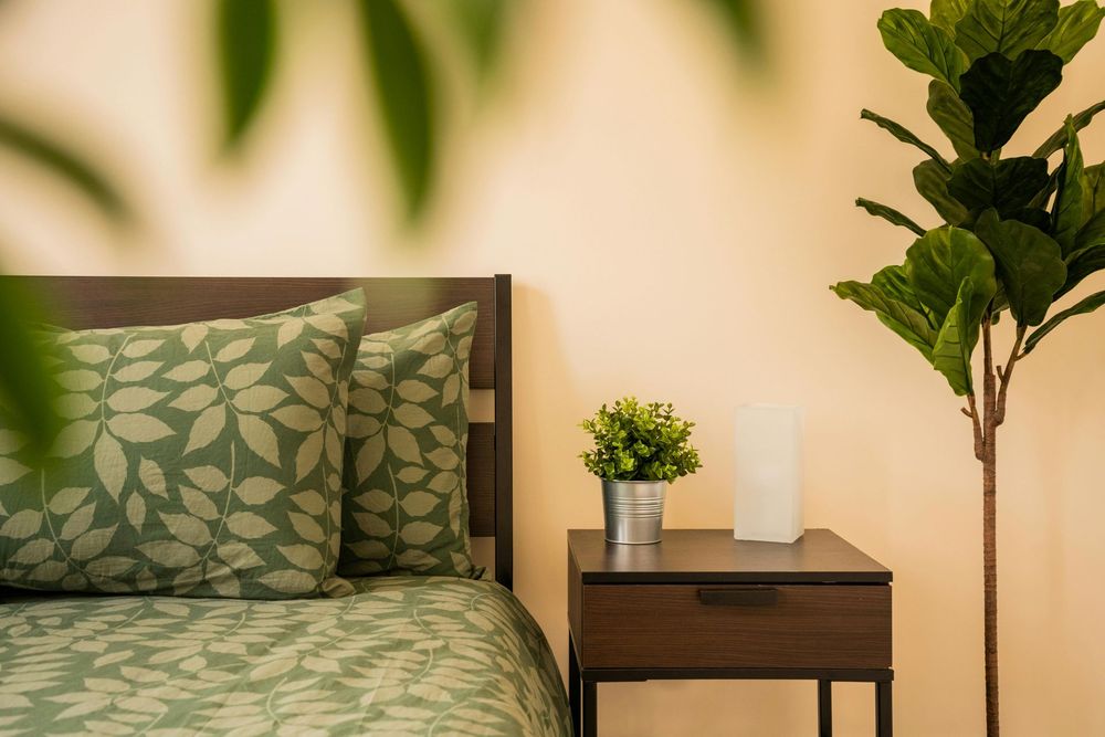 Bedroom with green leafy bedding, wooden nightstand, and potted plant against a beige wall.
