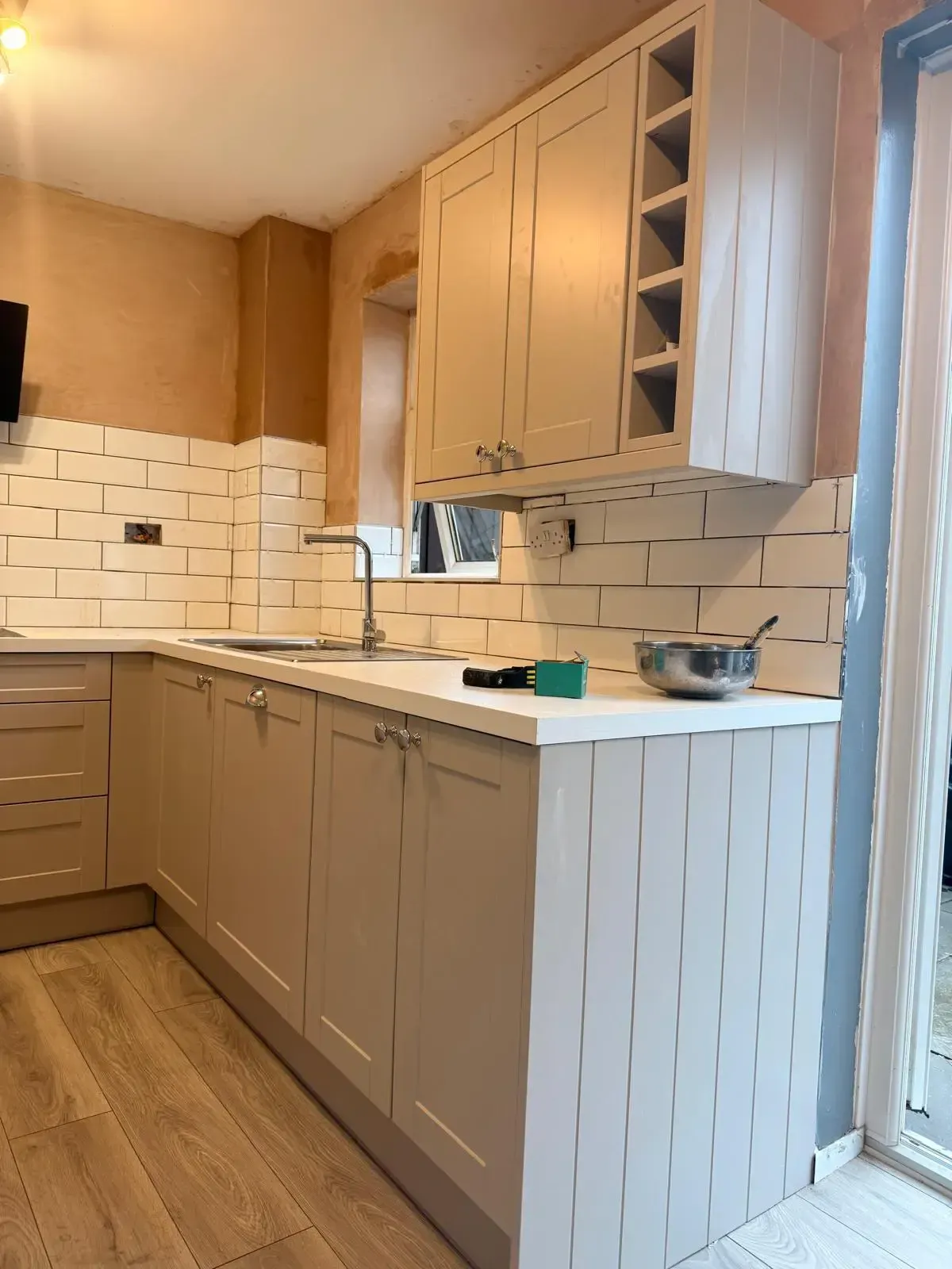 Small beige kitchen with white tile backsplash, wood cabinets, and a countertop near a window.