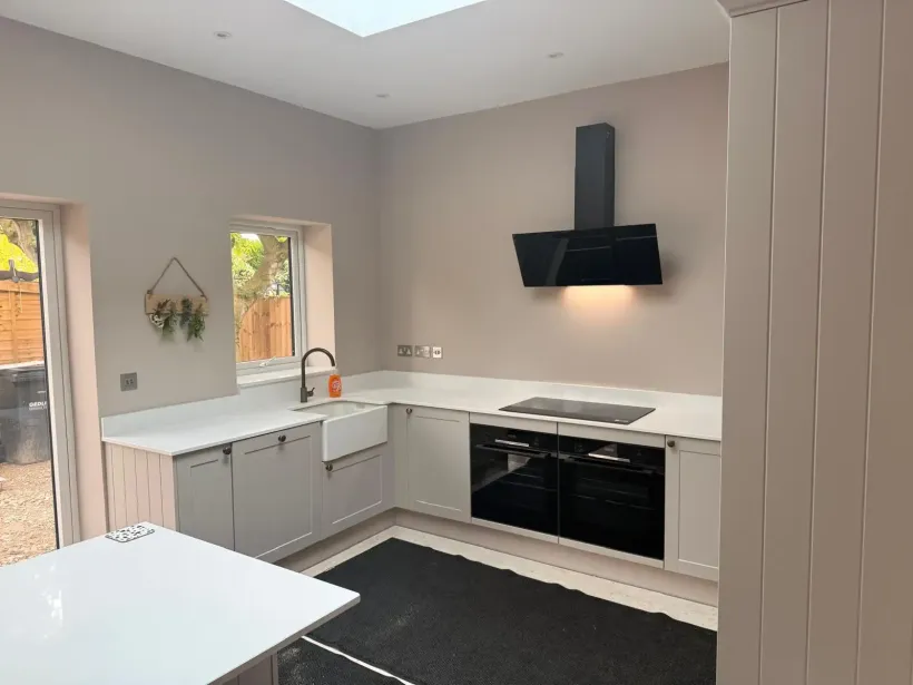 Modern L-shaped kitchen with light gray cabinets, white countertops, black oven and hood, and a farmhouse sink.