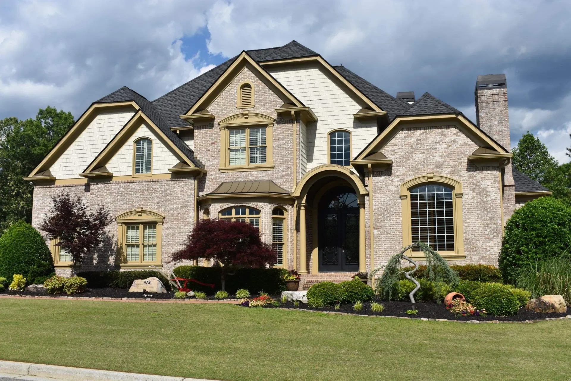 Two-story beige stucco house with dark roof and landscaped lawn under a cloudy sky.