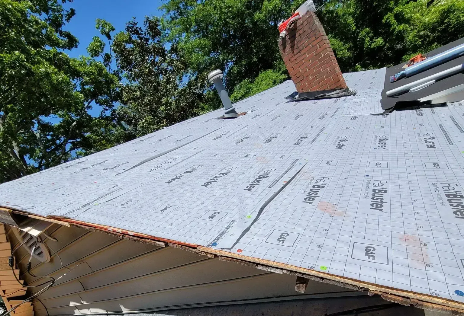 Rooftop with gray underlayment. Brick chimney, vent pipe, and trees in background.