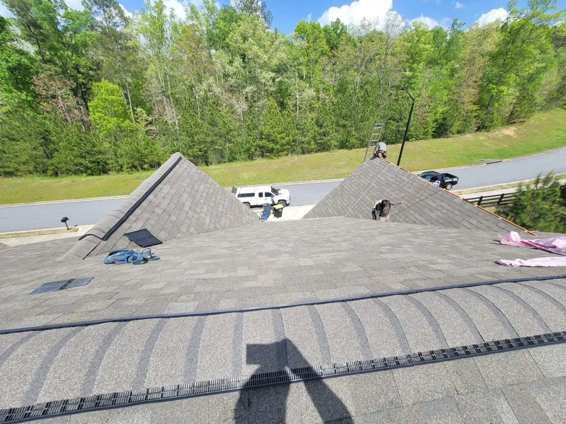 Workers on a gray shingle roof, two on the peaks, with a road and trees in the background.