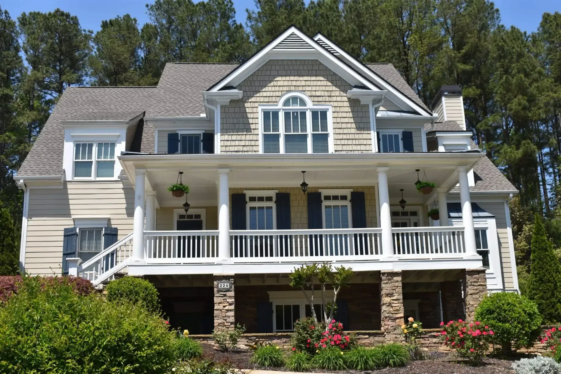 Two-story beige house with white porch, blue shutters, and stone foundation on a hill, surrounded by trees and landscaping.