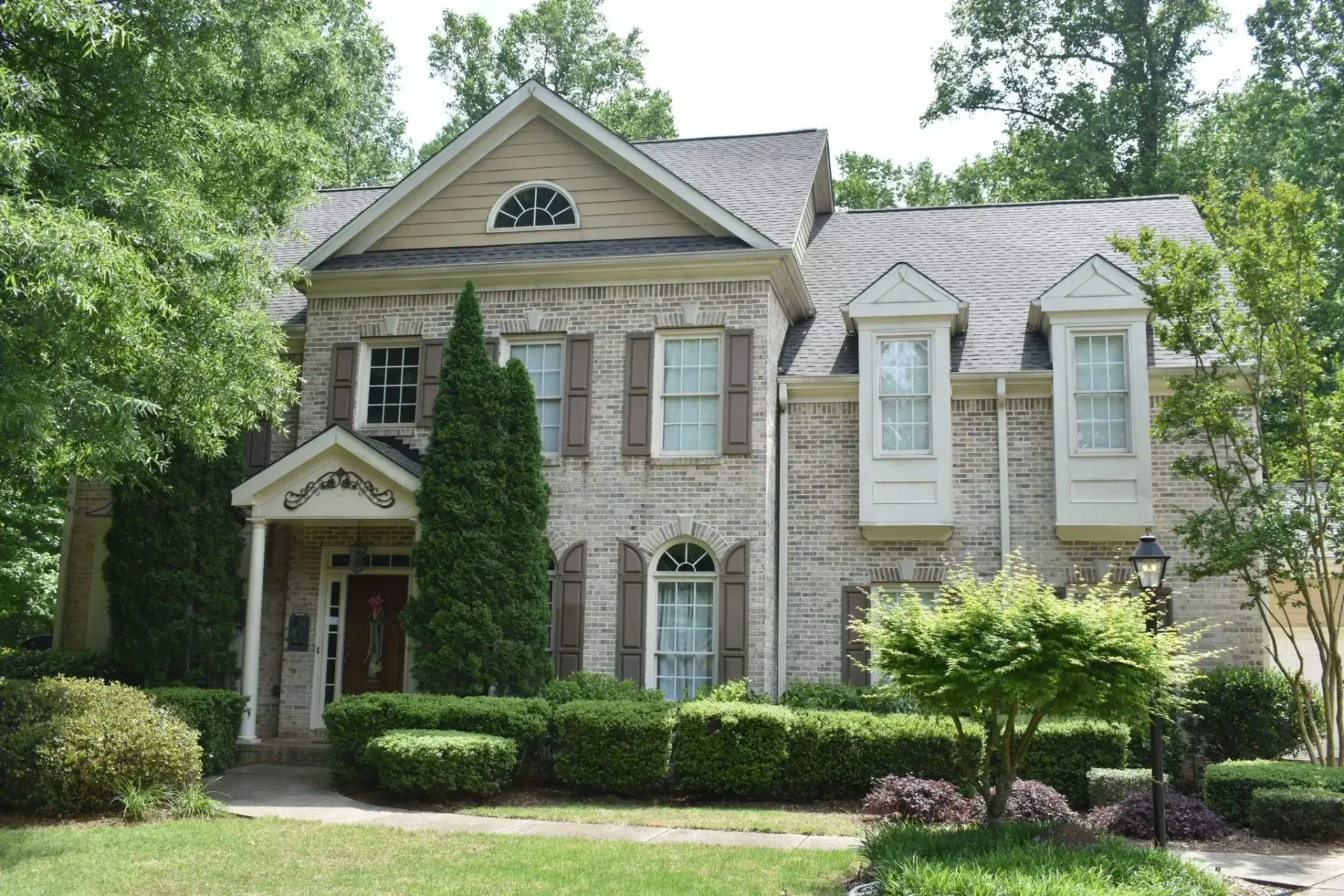 Two-story brick house with brown shutters, front porch, and dormer windows, surrounded by green trees and shrubs.