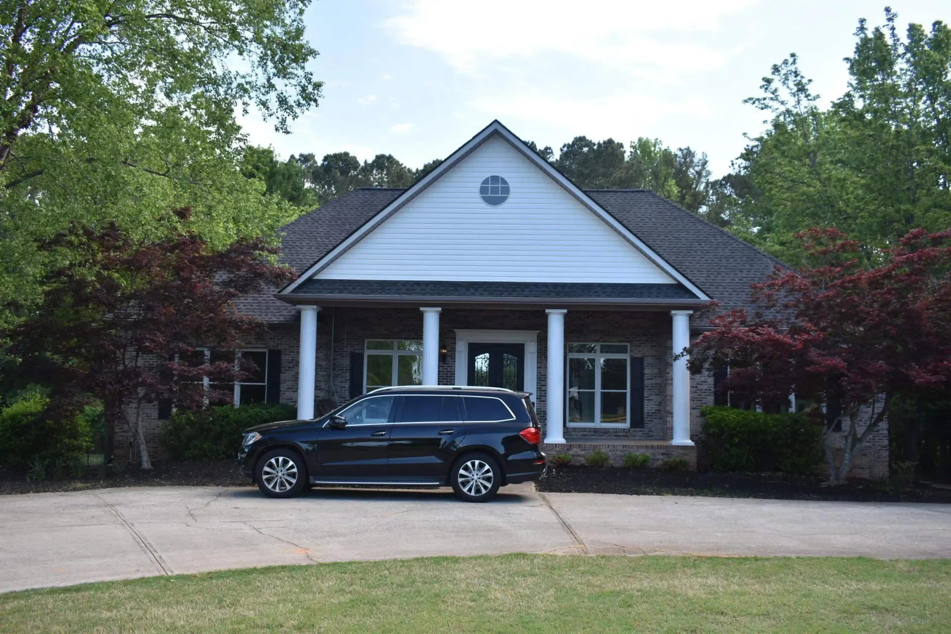 Black SUV parked in front of a house with white columns and a blue gable roof. Trees surround the house.