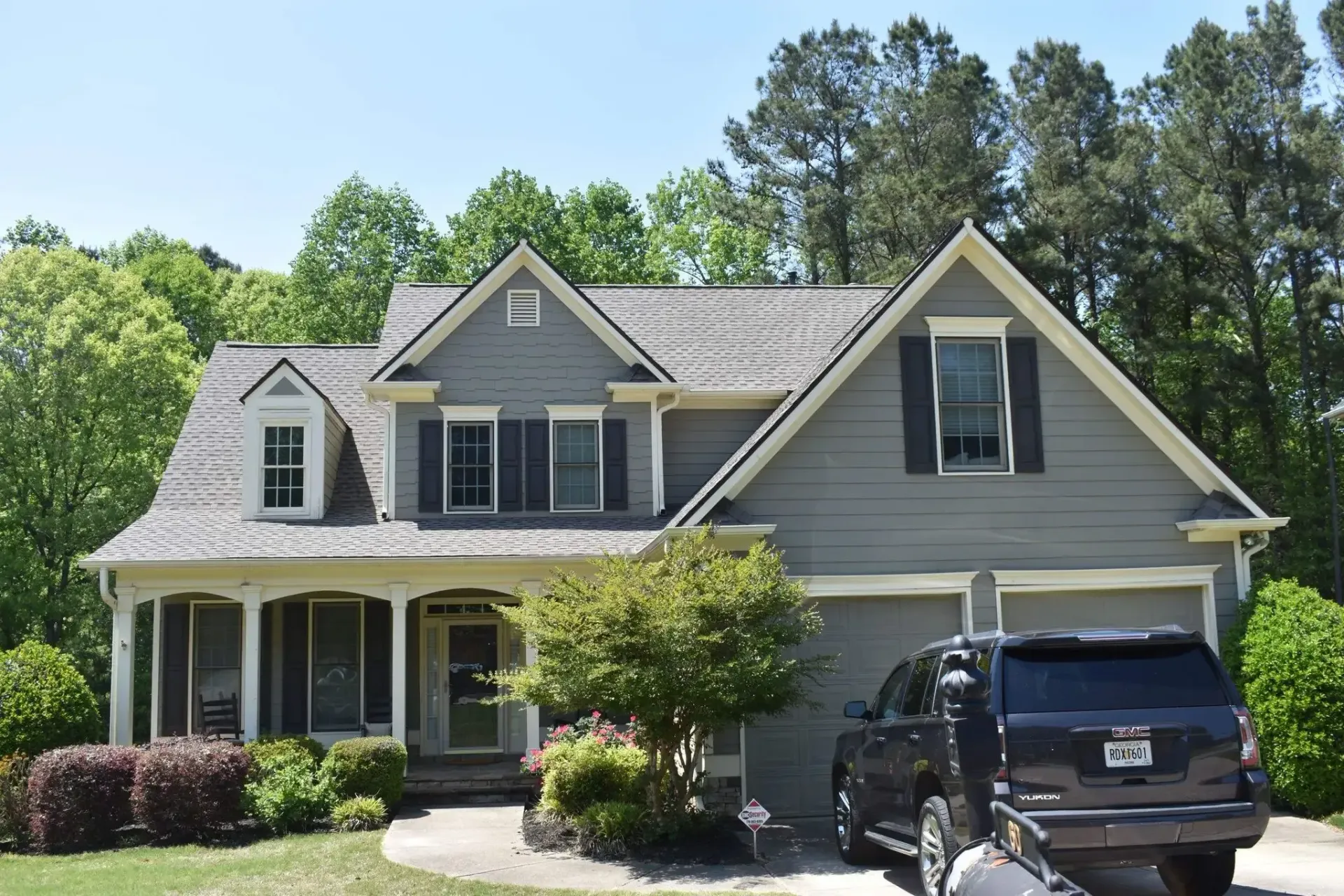 Gray house with shutters, garage, and black SUV in driveway. Green trees in background.