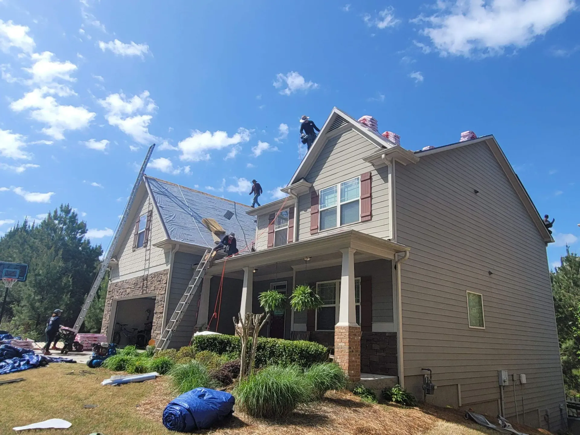 Roofers working on a two-story house with tan siding and a brick facade, under a partly cloudy sky.