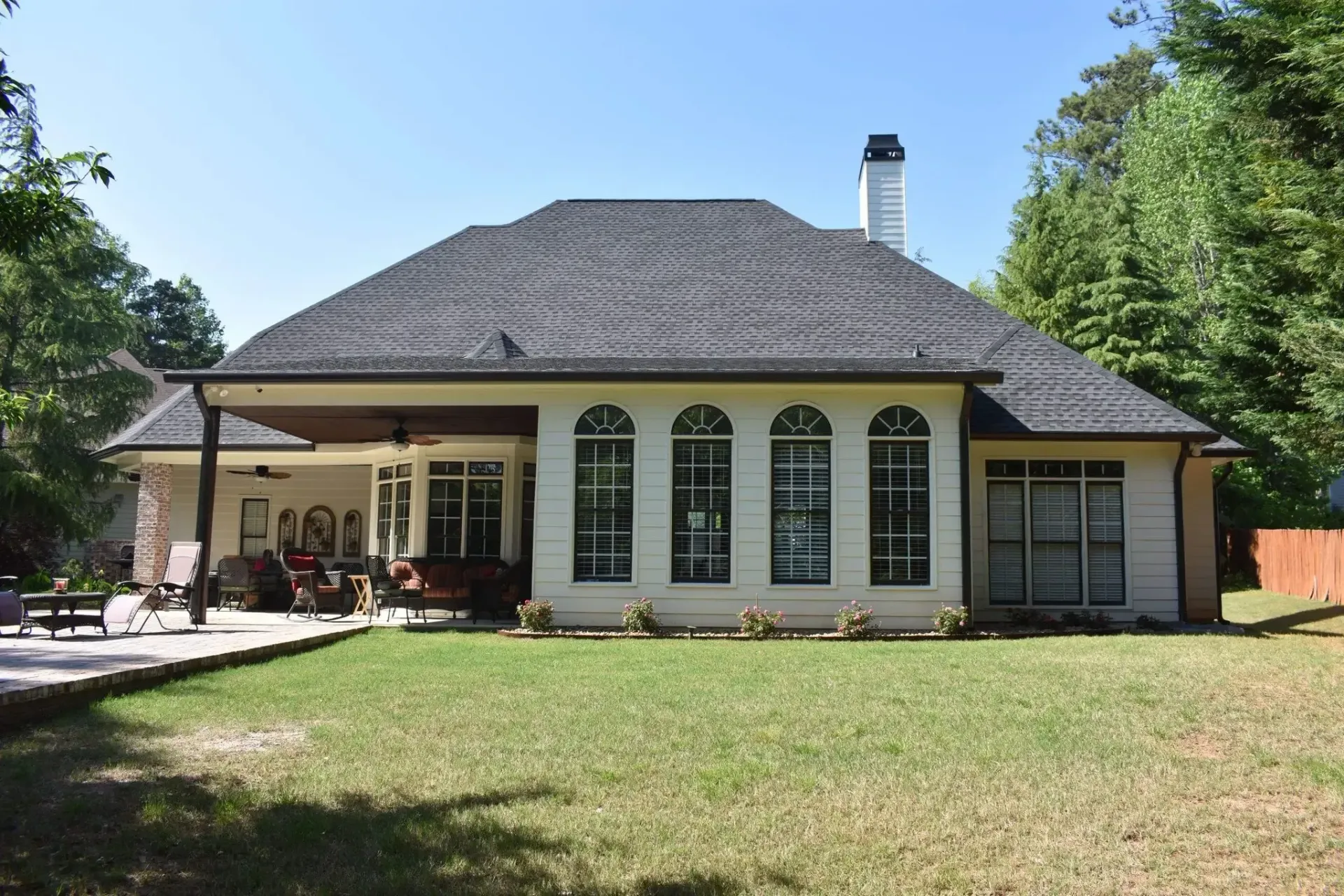 Back view of a light-colored house with a dark roof. Large windows, covered patio, and green lawn.