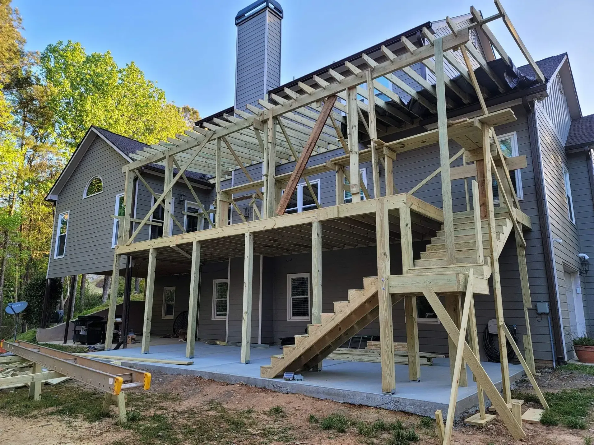 New wooden deck construction attached to a two-story gray house, with stairs, support beams, and a pergola.