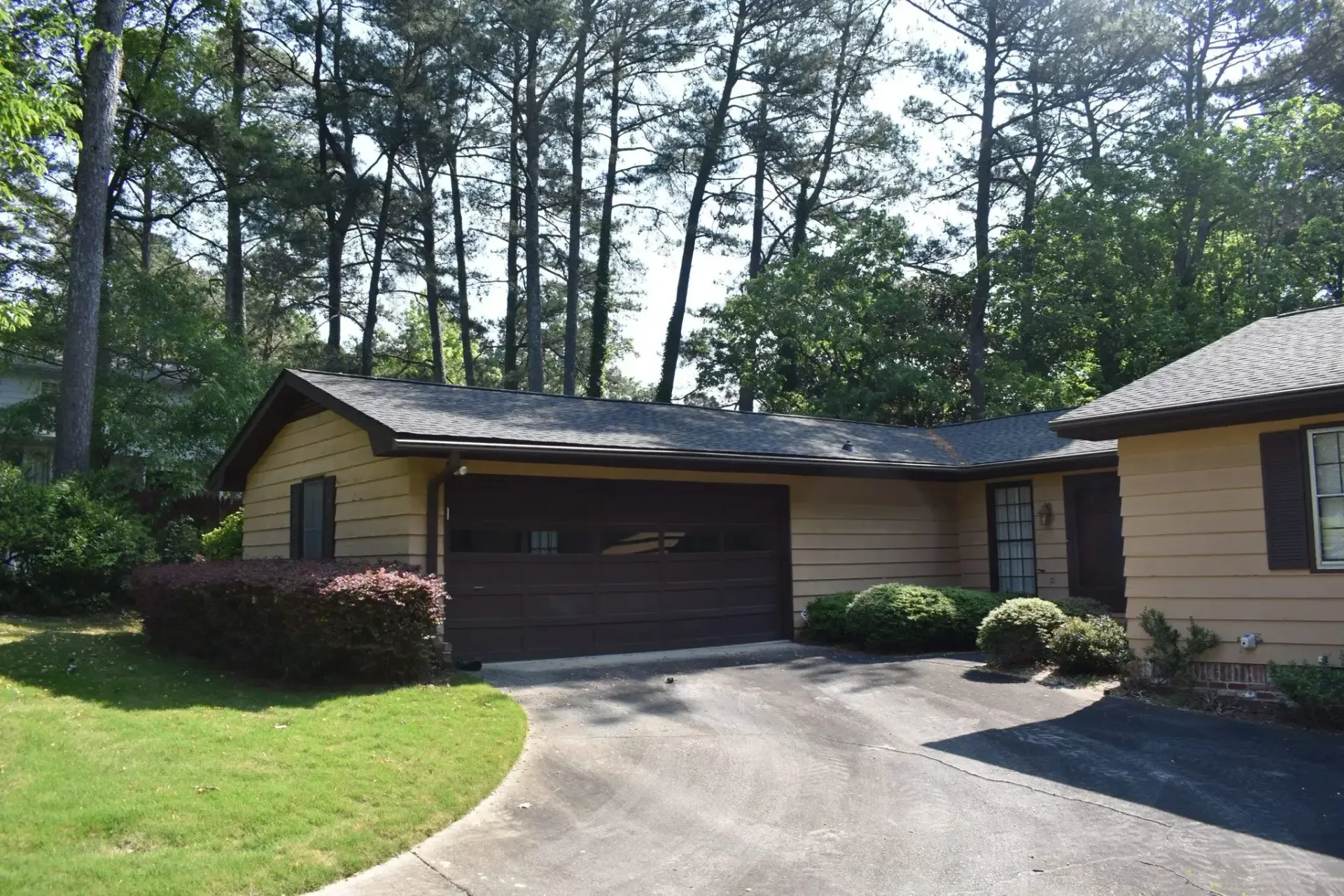Tan house with a brown garage door, dark roof, and a curved driveway, set among trees.