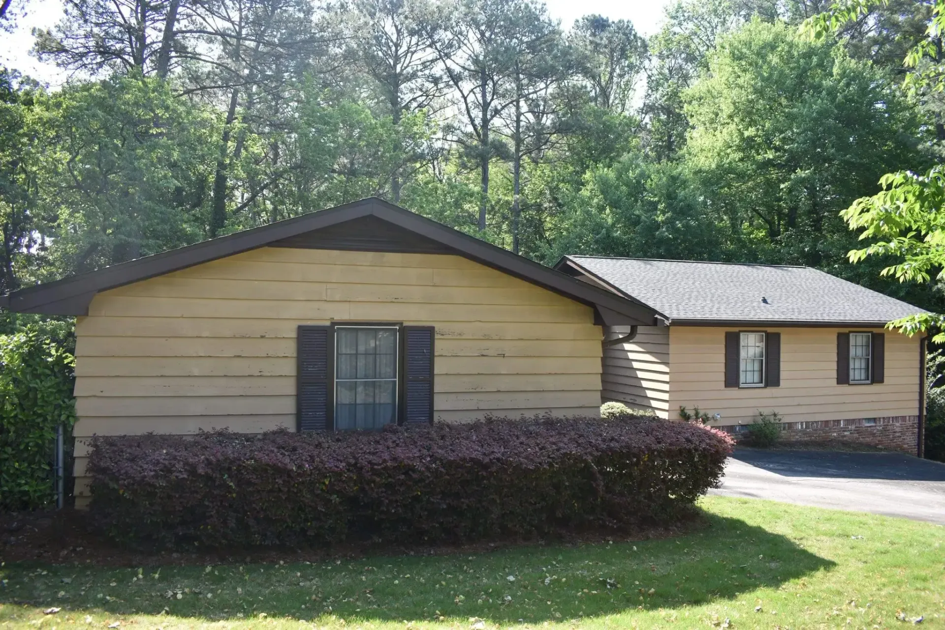 Beige house with dark shutters and roof, with a bush in front. Trees in background.