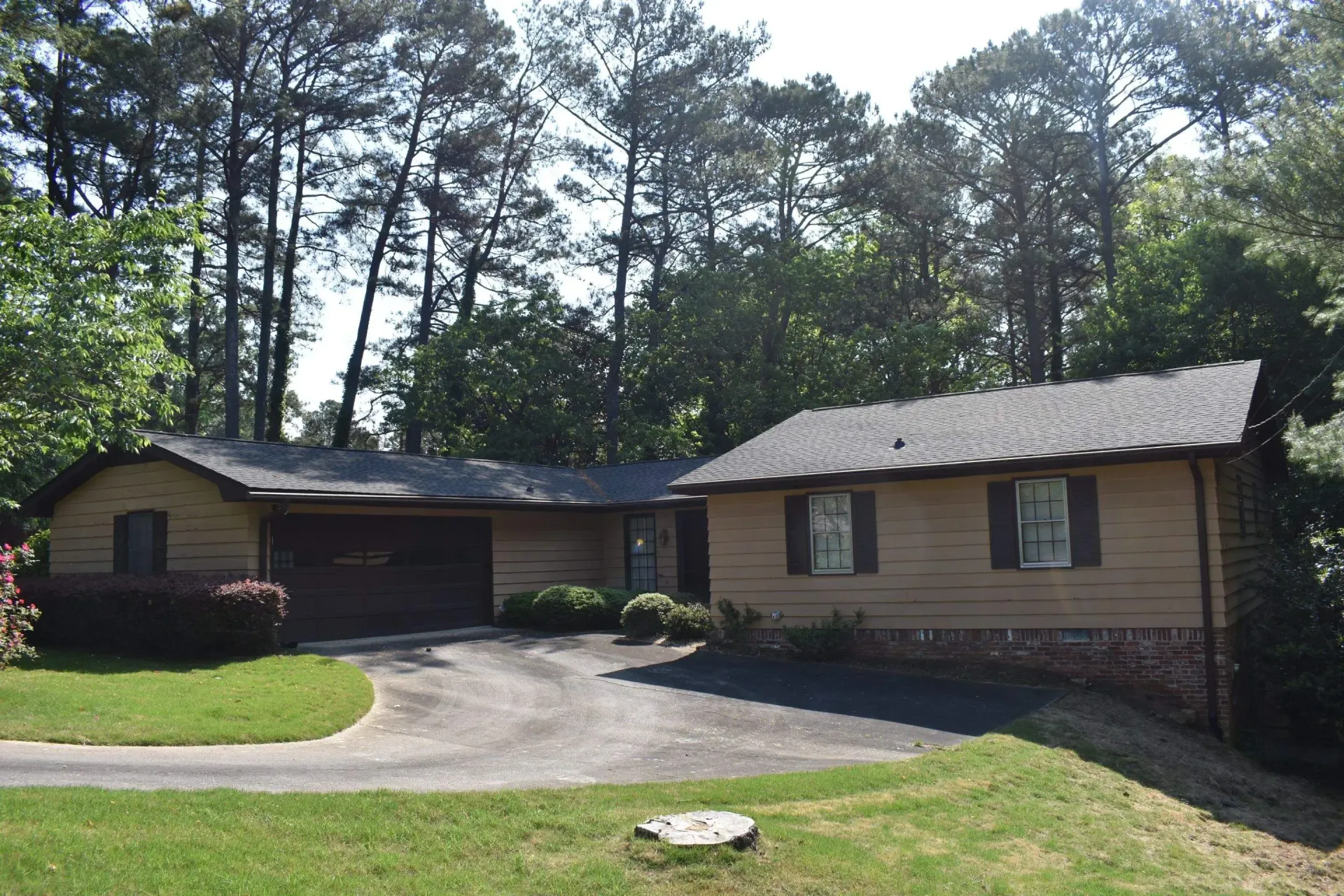 Tan ranch-style house with dark roof and garage, surrounded by trees. Driveway in front.